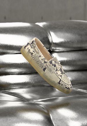 Madu mustriga tekstuuriga loafer-tüüpi king läbipaistva tallaga, hõbedase läikiva nupukohaga nahadiivani kohal hõljuv.