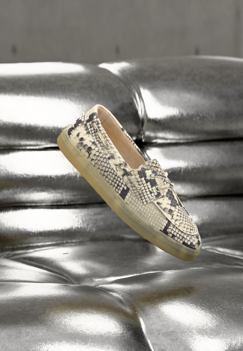 Παπούτσι loafer με υφή φιδιού και διάφανη σόλα που αιωρείται πάνω από γυαλιστερό ασημένιο καναπέ από δέρμα με ραφές.