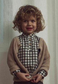 Barn som bär en beige stickad cardigan över en grå och vit rutig skjorta med knappar. Cardiganen har texturerade ärmar och träknappar.