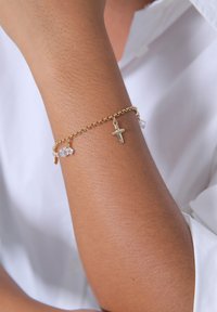 Guldkedja armbandskedja med ett litet korshänge och två klara ädelstenpendlar, vilande på ett solbrunt handled mot en vit bakgrund.