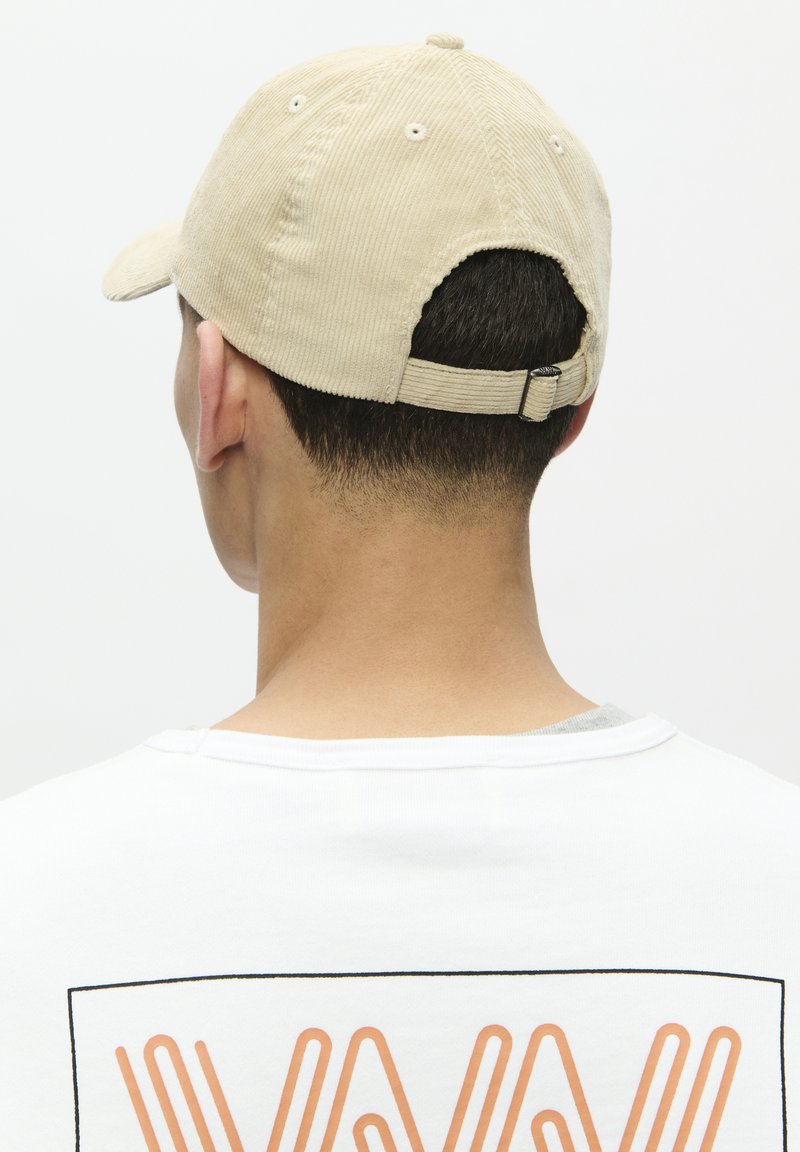 Beige corduroy cap with a curved brim, adjustable strap at the back, and visible stitching. Worn with a white shirt featuring an orange graphic.