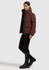 Donna che indossa un giubbotto imbottito rosso scuro, leggings neri e stivali al ginocchio neri, in piedi contro uno sfondo chiaro e semplice.