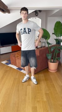 Siva majica s logotipom "GAP", črni uničeni kratki hlače in srebrni športni copati. Ozadje vključuje rjavo sedežno garnituro in sobne rastline.