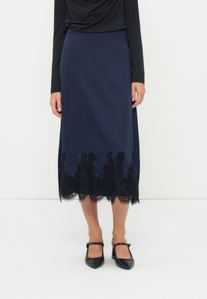 Frau steht und trägt einen navyblauen Midirock mit schwarzer Spitzenverzierung am Saum, ein schwarzes Oberteil mit langen Ärmeln und schwarze Mary-Jane-Schuhe.