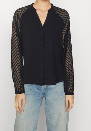 Femme portant un chemisier noir boutonné avec des manches longues transparentes à motifs et un jean bleu clair, debout devant un fond uni.