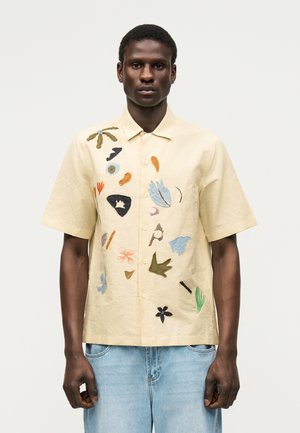 Mees, kes kannab beeži lühikese varrukaga nööbitavat särki, millel on värvikad abstraktsed tikitud kujundid, ja helesiniseid teksaseid ühtlasel taustal.