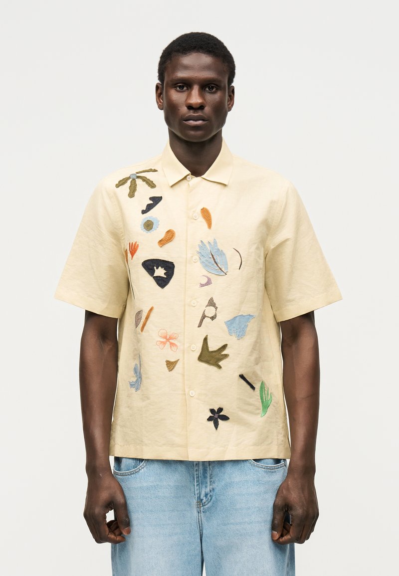 Άνδρας με μπεζ κοντομάνικο πουκάμισο με πολύχρωμα αφηρημένα κεντημένα σχέδια και ανοιχτό μπλε τζιν σε μονόχρωμο φόντο.