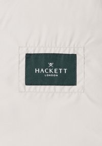 Étiquette en tissu vert foncé cousue sur un matériau beige clair, affichant le nom de la marque "Hackett London" et un logo de rames croisées en texte blanc.