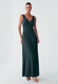 Žena stojí sebavedome v priliehavých tmavozelených maxi šatách, na sebe má elegantné zlaté doplnky a sandále na vysokom opätku v nude farbe, pričom pozadie je neutrálné.