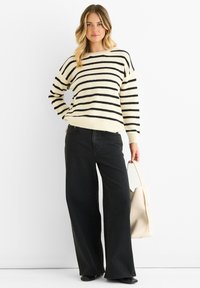 Gestreifter Pullover in Creme und Schwarz, kombiniert mit hochtaillierten, weiten schwarzen Jeans. Akzentuiert mit einer hellen Handtasche.