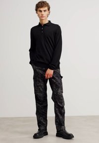 Camisa polo de manga longa preta combinada com calças cargo de camuflagem escuras e botas pretas robustas. Botões visíveis na camisa polo e bolsos laterais nas calças.