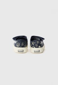 Sapatos de lona azul-marinho com padrão de conchas brancas e estrelas-do-mar, apresentando uma tira de velcro e sola de borracha em tom creme. Marca Superga visível.