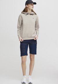 Hoodie beige clair avec une poche avant et un cordon de serrage ajustable, associé à un short cargo navy avec une poche latérale et un bouton. Baskets blanches.