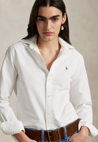 Kobieta z długimi ciemnymi włosami nosi białą koszulę zapinaną na guziki z podwiniętymi mankietami, brązowy pasek, niebieskie dżinsy oraz wiszącą kolczyk z muszelką.