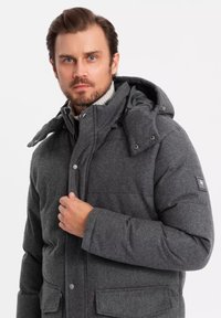 Giubbotto puffo grigio con cappuccio, realizzato in un tessuto texturizzato, con chiusura a scatto e due tasche frontali. Patch del logo ben visibile sulla manica.