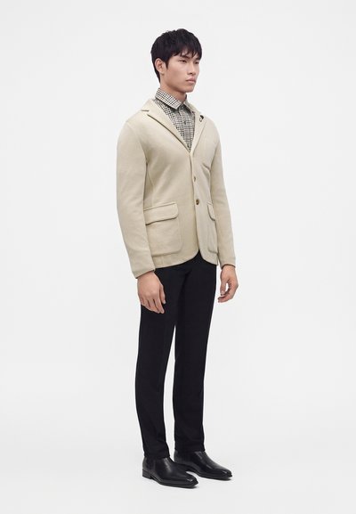 Homme portant un blazer beige, une chemise à carreaux, un pantalon noir et des chaussures habillées noires, debout sur un fond blanc uni.