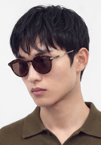 Jeune homme portant des lunettes de soleil rondes en écaille et une chemise à col vert olive, regardant légèrement sur le côté sur un fond uni.
