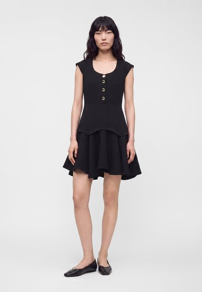 Femme debout portant une robe noire sans manches avec des boutons dorés et une jupe évasée, associée à des chaussures plates noires.