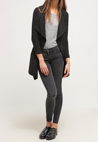 Svart öppen kappa, grå topp, åtsittande svarta jeans och svarta skor. Kappan har en draperad krage och mjuk textur.