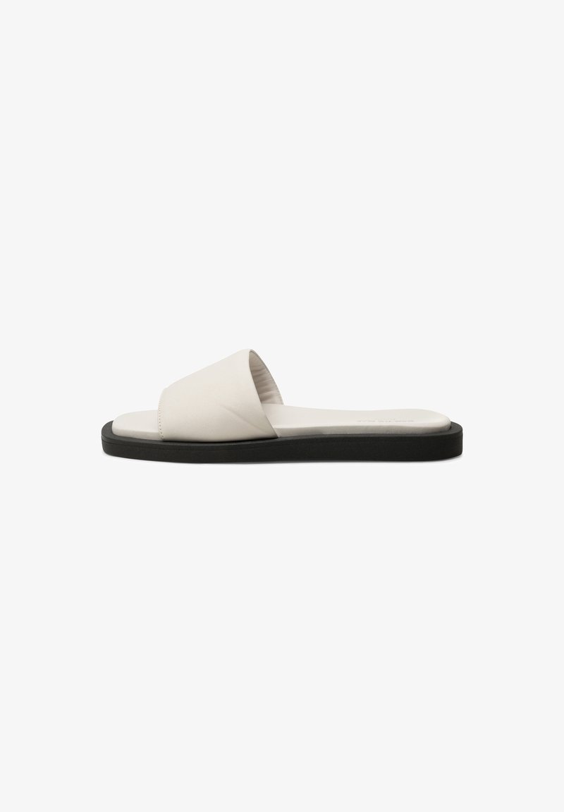 Sandales plates à glissière avec une tige en similicuir crème, un design à large sangle, et une semelle en caoutchouc noire, présentant une texture lisse et un style minimaliste.