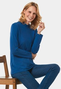 Blaue Langarm-Turtleneck-Oberteile aus weichem Stoff, mit lockerem Schnitt und glatter Textur, kombiniert mit dunkelblauen Jeans. Sie sitzt auf einem Holzstuhl.