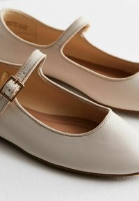Zapatos Mary Jane de cuero beige con punta redonda, correa con hebilla dorada y detalle de borde cosido. Textura suave.