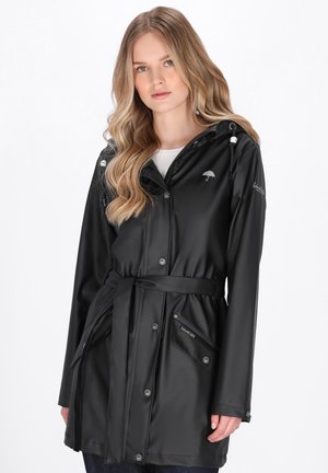 Fekete, deréknál megkötős esőkabátot viselő nő, kapucnival, gombos és cipzáras záródással, egyszerű fehér háttér előtt állva.