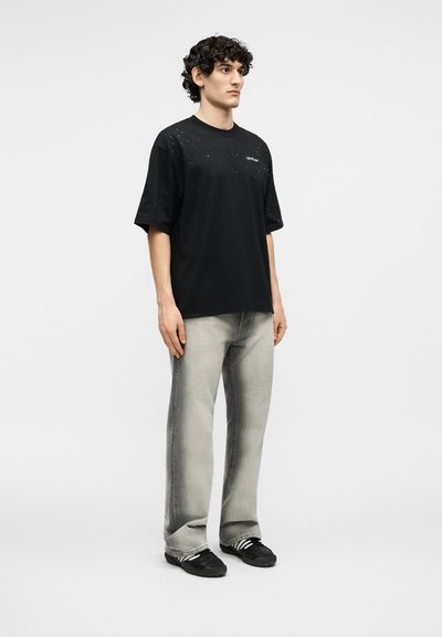 Νέος άνδρας όρθιος, φοράει μια μαύρη υπερμεγέθη μπλούζα με στίγματα, ξεβαμμένο γκρι τζιν με ίσια πόδια και μαύρα αθλητικά παπούτσια με λευκές ρίγες.