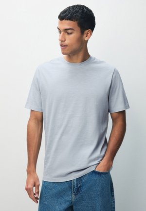 Camiseta de manga corta gris claro hecha de una tela suave, con un cuello redondo clásico y un corte holgado. Combinada con jeans de mezclilla azul.