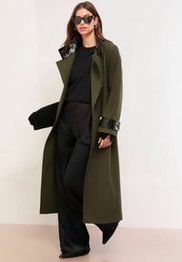 Długi oliwkowozielony płaszcz z czarnym błyszczącym kołnierzem, zestawiony z czarnym topem i spodniami o szerokich nogawkach, z koronkowymi zdobieniami u dołu. Czarne spiczaste buty.