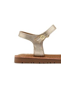 Sandalia metálica de bronce con una sola correa, plantilla de ante color marrón y suela de goma texturizada con recortes triangulares.