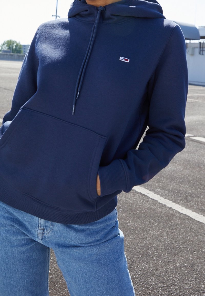 Personne portant un sweat à capuche bleu marine avec poche avant et un jean bleu clair, debout à l'extérieur sur une surface en asphalte.