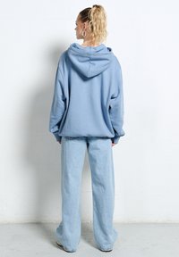 Kobieta z blond kucykiem, ubrana w za dużą jasnoniebieską bluzę z kapturem i szerokie jasnoniebieskie dżinsy, stoi twarzą do białej ściany.