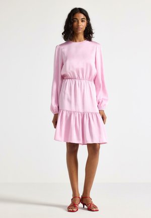 Kvinde iført en lyserød langærmet kjole med tyllet talje og lagdelt nederdel, kombineret med røde sandaler med lav hæl og remme.