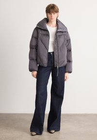 Grå polstret jakke med høy krage og glidelås, sammen med mørkeblå vide jeans. Jevn stofftekstur og geometriske stikningsdetaljer.