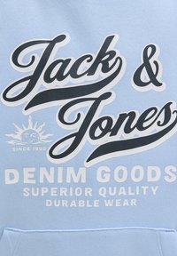 Hoodie bleu clair avec un texte audacieux en noir et blanc : "Jack & Jones," "DENIM GOODS," "QUALITÉ SUPÉRIEURE," et "USAGE DURABLE." Graphique de soleil inclus.