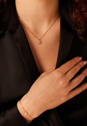 Gouden ketting en armband met een roze bloemenhanger versierd met meerdere stenen, gedragen tegen een zwarte stoffen achtergrond.