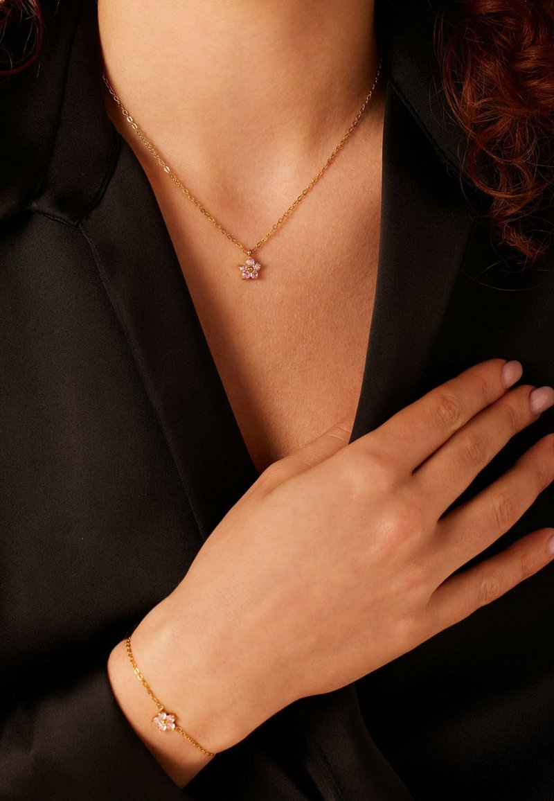Gouden ketting en armband met een roze bloemenhanger versierd met meerdere stenen, gedragen tegen een zwarte stoffen achtergrond.