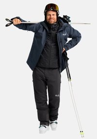 Skijack in donkerblauw, geïsoleerd zwart vest eronder, zwarte skibroek, witte skischoenen, met twee witte skistokken in de hand, en een skibril op.