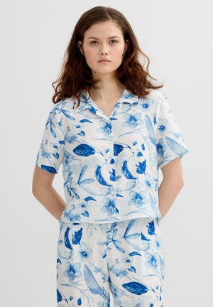 Mujer joven con camisa de manga corta y pantalones a juego con estampado floral azul sobre fondo blanco, de pie con las manos detrás de la espalda.