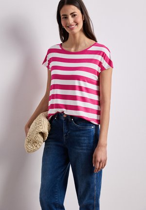 Femme souriante portant un t-shirt à manches courtes rayé rose et blanc et un jean bleu, tenant un chapeau en tricot beige sur un fond uni.