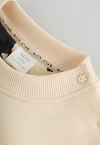Sudadera de color beige claro con cuello de canalé, textura suave y etiqueta distintiva que presenta el logo "NEXT baby" y "milk club".