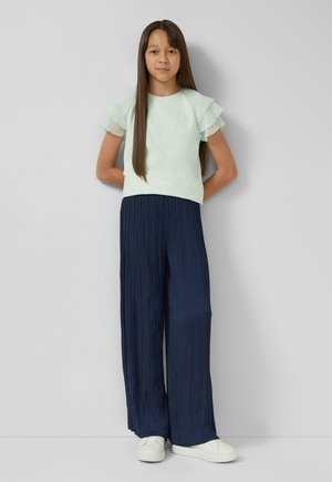 Hellgrünes Oberteil mit geschichteten, gerafften Ärmeln, kombiniert mit marineblauen, plissierten Hosen mit weitem Bein. Weiße Sneakers vervollständigen das Outfit.