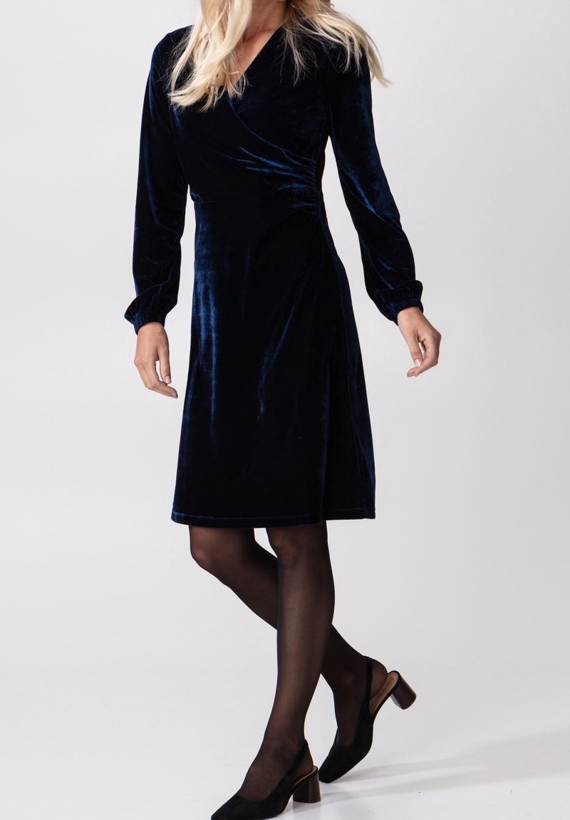 Femme portant une robe en velours bleu foncé à manches longues, longueur genou, collants noirs transparents et chaussures slingback noires à talons blocs.