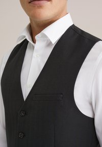 Melns hounds-tooth vestes ar izgrieztu uzvalku, ar divām brūnām pogām un krūšu kabatu virs baltas pogājamas krekla.