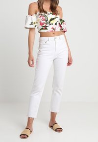 Kvinna iförd blommig crop top med avskuren axel, högmidjade vita jeans med råa kanter, och beige vävda sandaler stående framför en neutral bakgrund.