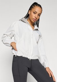 Casaco leve branco com fecho de correr e gola alta, com punhos elásticos e bainha com cordão, usado com leggings pretas com costuras laterais.