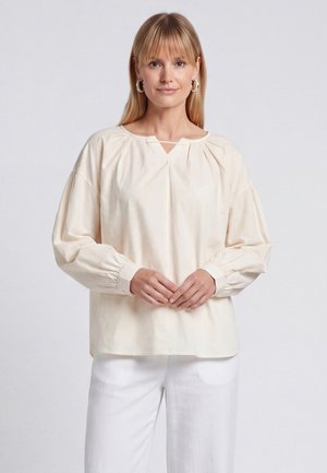 Blonde Frau mit Creolen, die eine hellbeige Bluse mit langen Ärmeln und gesammelten Manschetten sowie weiße Hose trägt, stehend vor einem schlichten Hintergrund.