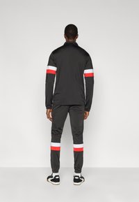 Μαύρο αθλητικό μπουφάν και παντελόνι με λευκές και κόκκινες ρίγες στα μανίκια και στους αστραγάλους. Λείο ύφασμα, κανονική εφαρμογή, μινιμαλιστικός σχεδιασμός.