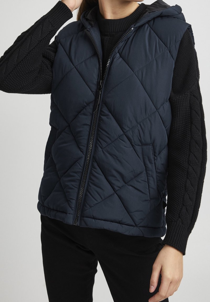 Femme portant une veste sans manches matelassée bleu marine avec capuche par-dessus un pull en maille torsadée noir, ajustant ses cheveux d'une main.